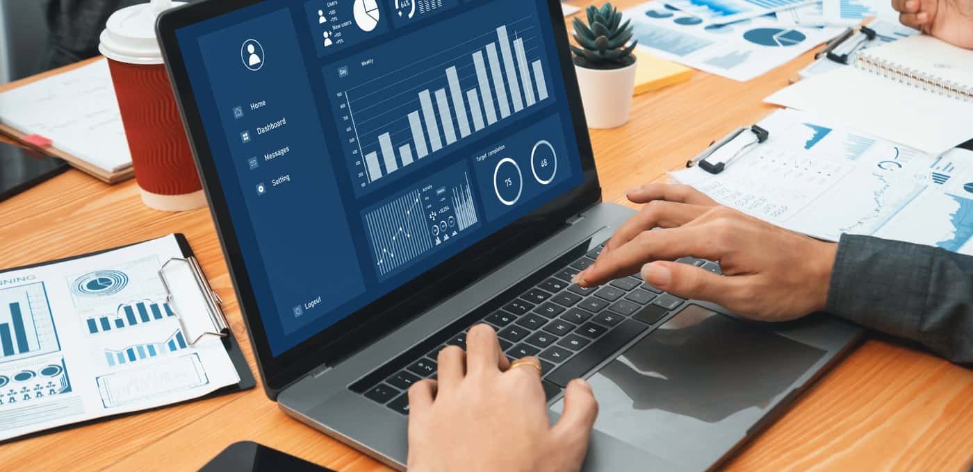 Hands typing on a laptop showing a blue analytics dashboard, with printed performance charts and a coffee on the desk beside it.